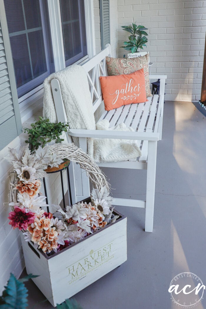A white wooden bench on a simple porch decorated for fall, featuring a white throw blanket, two decorative pillows, and a rustic wooden crate filled with an autumn-themed floral wreath.