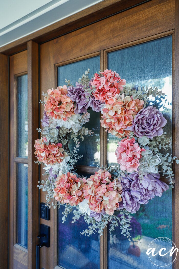 A floral wreath of peach, purple, and white hydrangea blossoms with eucalyptus greenery hanging on a wooden front door, perfect for simple porch decor.