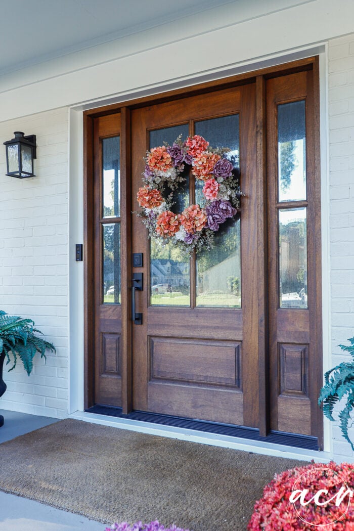 A dark wood front door with glass panels and a wreath of orange and purple flowers, set in a white brick wall for simple fall decor.
