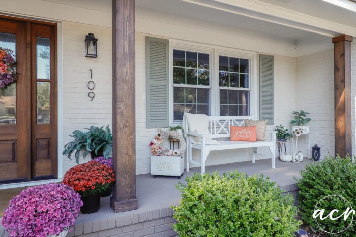 A front porch with a white bench, harvest-themed fall decor, potted mums, and a wooden front door.