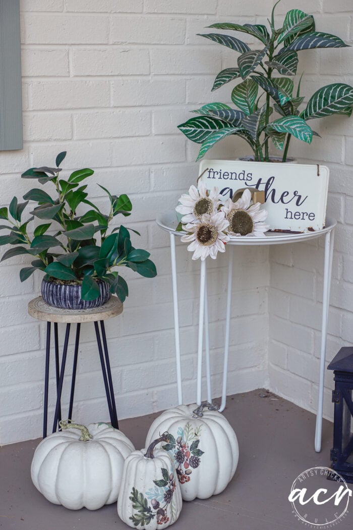 Two potted plants on metal stands, a sign that reads friends gather here, three faux sunflowers, and three decorative white pumpkins create simple fall decor on a porch.