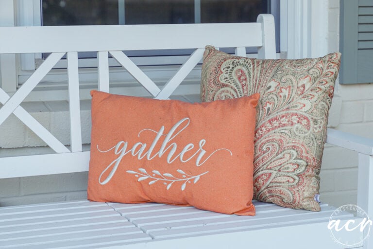 Two decorative pillows on a white wooden bench for simple fall decor; one is orange with the word gather embroidered on it, and the other has a tan and orange paisley pattern.