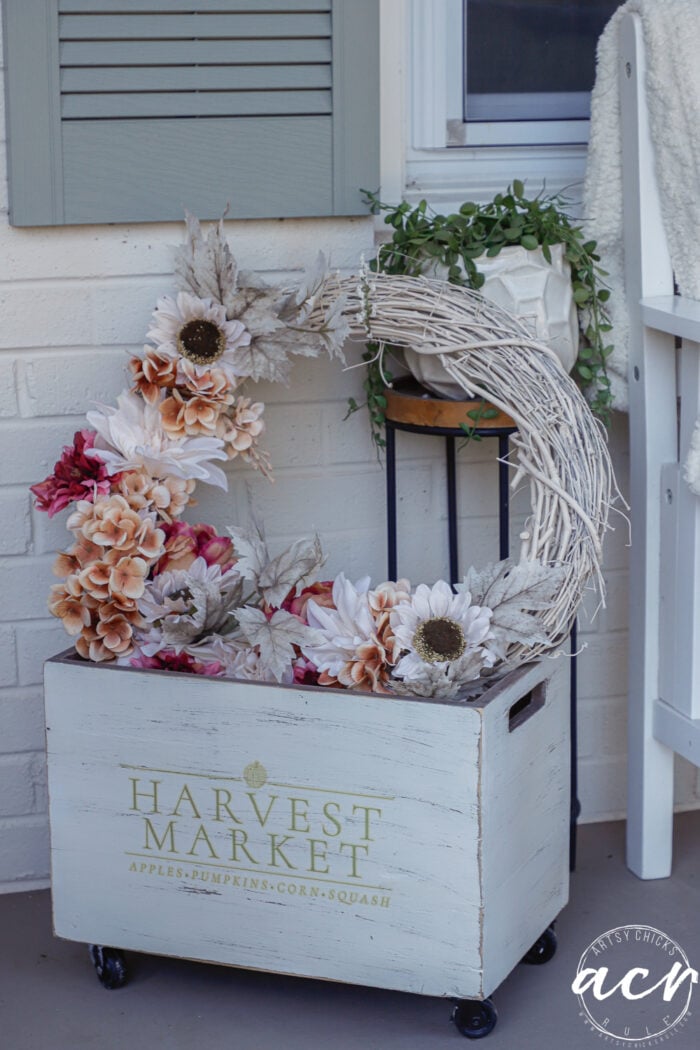 A white wooden crate on wheels labeled Harvest Market, filled with an autumn-themed floral wreath, creates simple fall decor for a porch.
