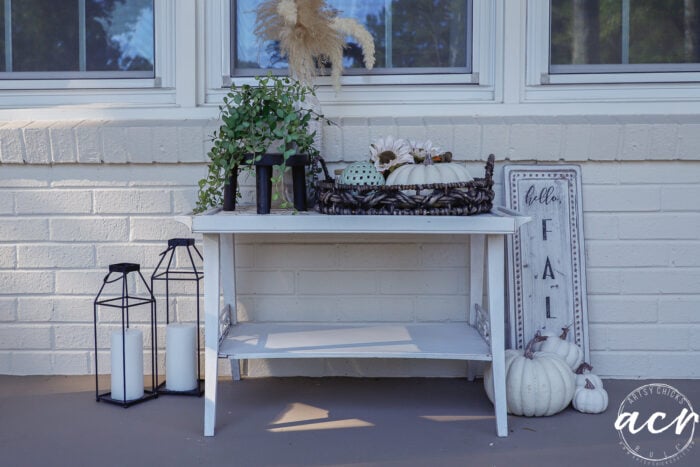 A white porch table styled with simple fall decor, featuring a potted plant, a woven basket of pumpkins, and a hello FALL sign, accented by lanterns and additional pumpkins on the floor.