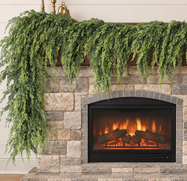 A lush faux cedar garland draped over a stone fireplace mantel with a lit electric fire, a standout among Christmas favorites.