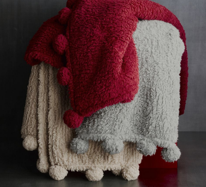 Three plush, textured throw blankets in red, gray, and beige, each featuring pom-pom trim along the edge as holiday favorites.